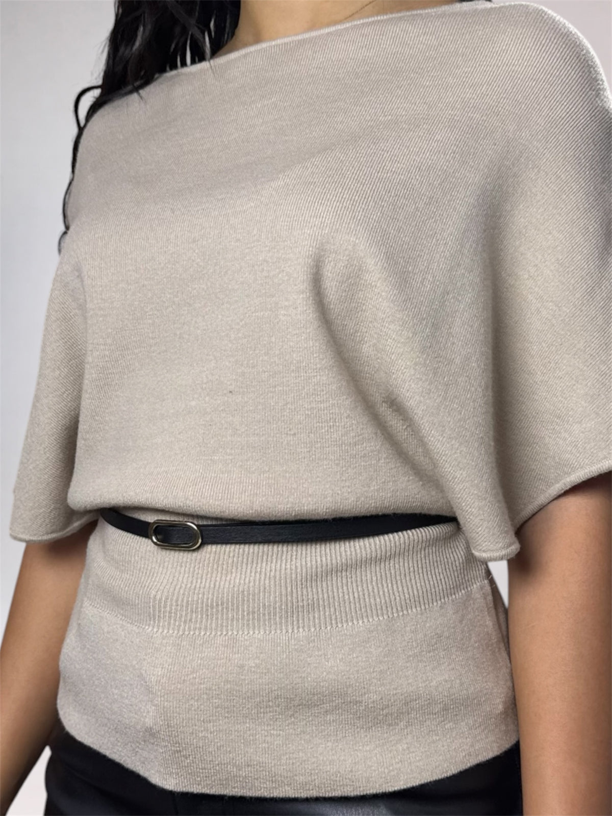 A close-up of a person wearing a light beige knit top with a wide boat neckline and short wide sleeves, cinched at the waist by a thin black belt with a silver oval buckle.