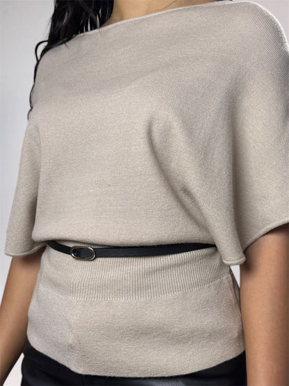 A close-up of a person wearing a light beige knit top with a wide boat neckline and short wide sleeves, cinched at the waist by a thin black belt with a silver oval buckle.