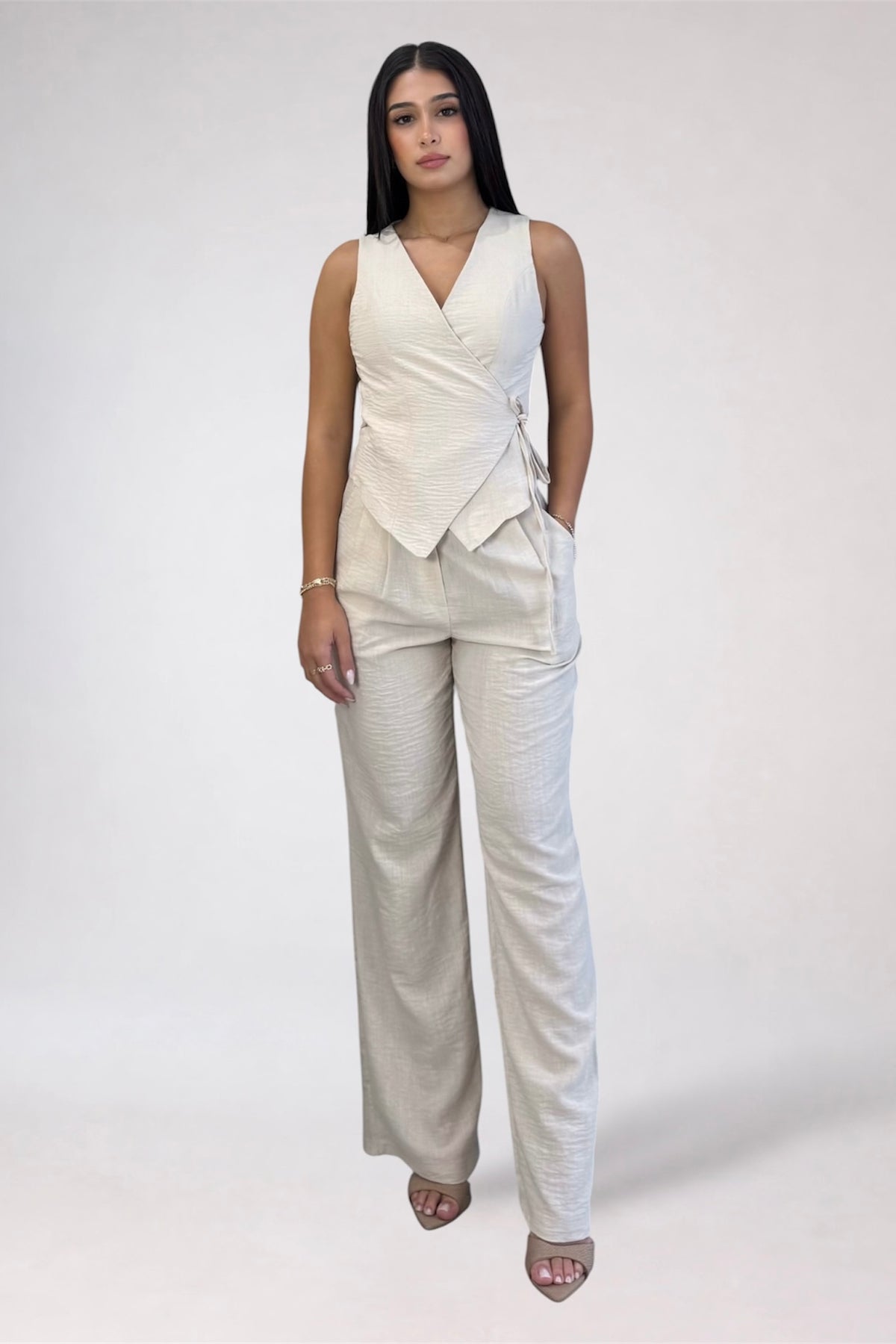 A woman modeling a sleeveless wrap-style top and matching wide-leg trousers in a light cream or off-white color, standing against a plain light gray studio background.