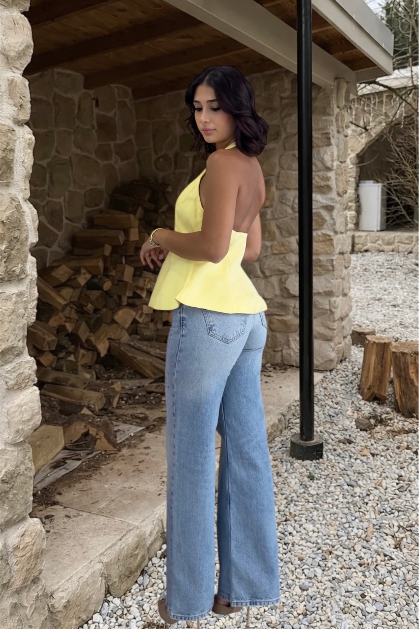 Woman in yellow top and blue jeans standing near a stone wall with stacked firewood.