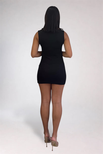 A woman viewed from behind wearing a sleeveless black mini dress and nude high heels, standing against a plain light gray studio background.