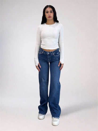 A model stands against a light gray studio background wearing a fitted white long-sleeve crop top, blue high-waisted straight-leg jeans with subtle distressing, and white sneakers.