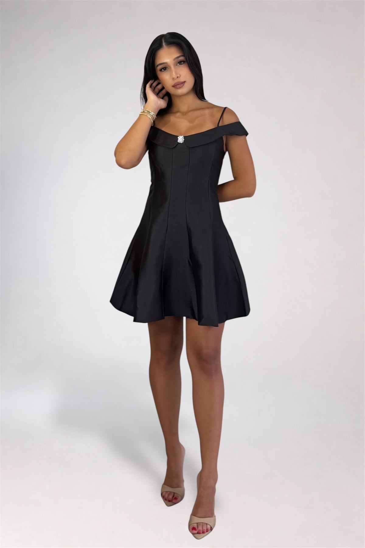A woman modeling a short black off-the-shoulder dress with a structured bodice and flared skirt, paired with nude high-heeled sandals, against a light gray studio background.