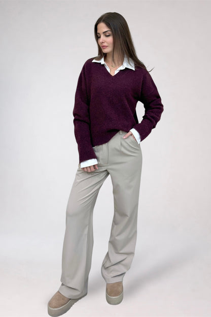 Woman wearing a purple sweater, white shirt, and beige pants on a plain background