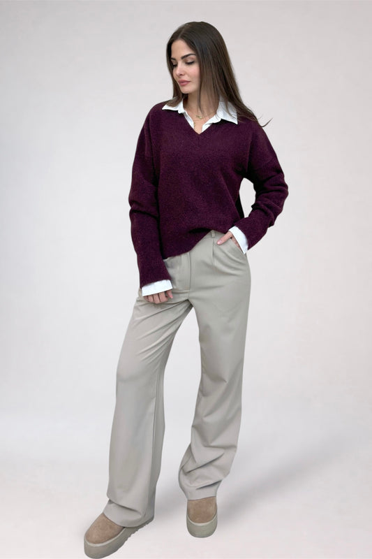 Woman wearing a purple sweater, white shirt, and beige pants on a plain background