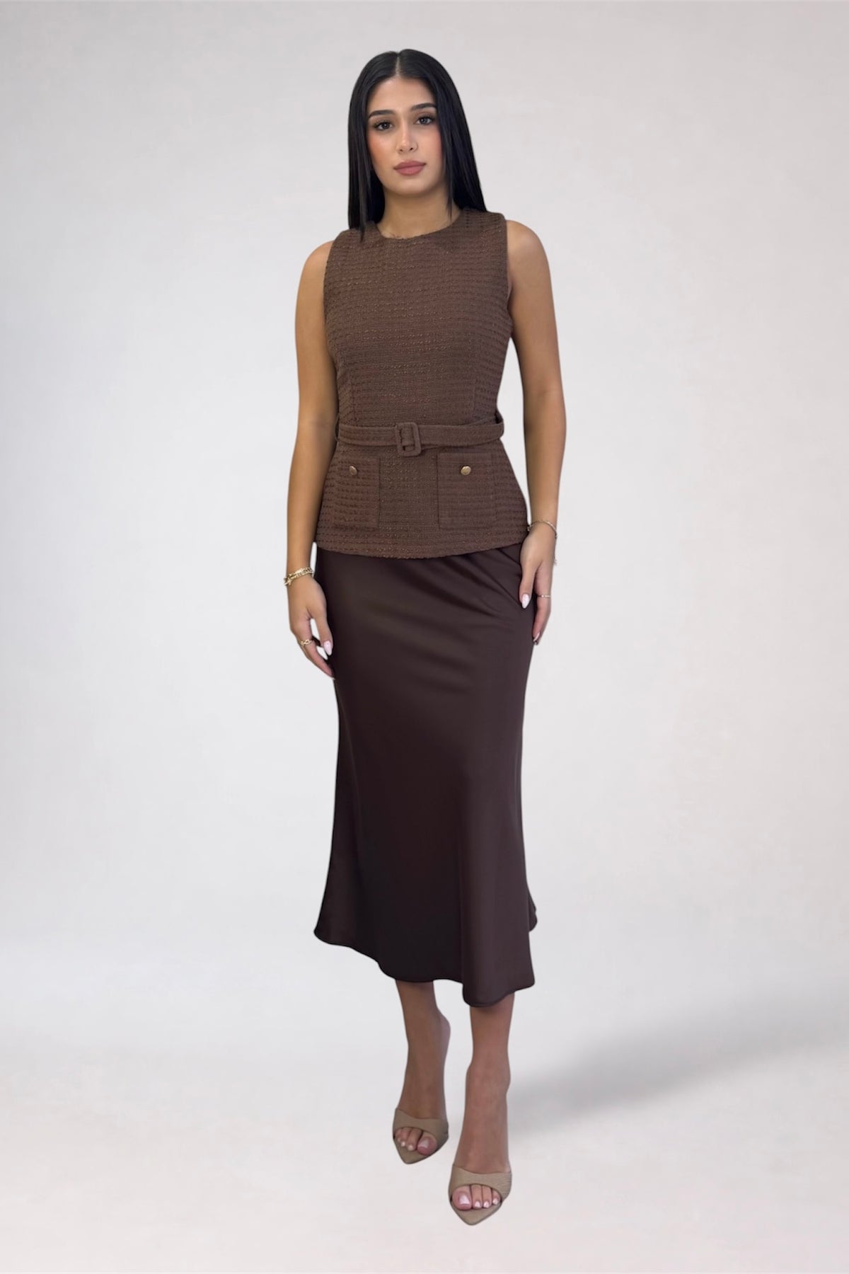 A woman modeling a sleeveless brown textured top with a matching belt and a dark brown midi skirt, standing against a light gray studio background.