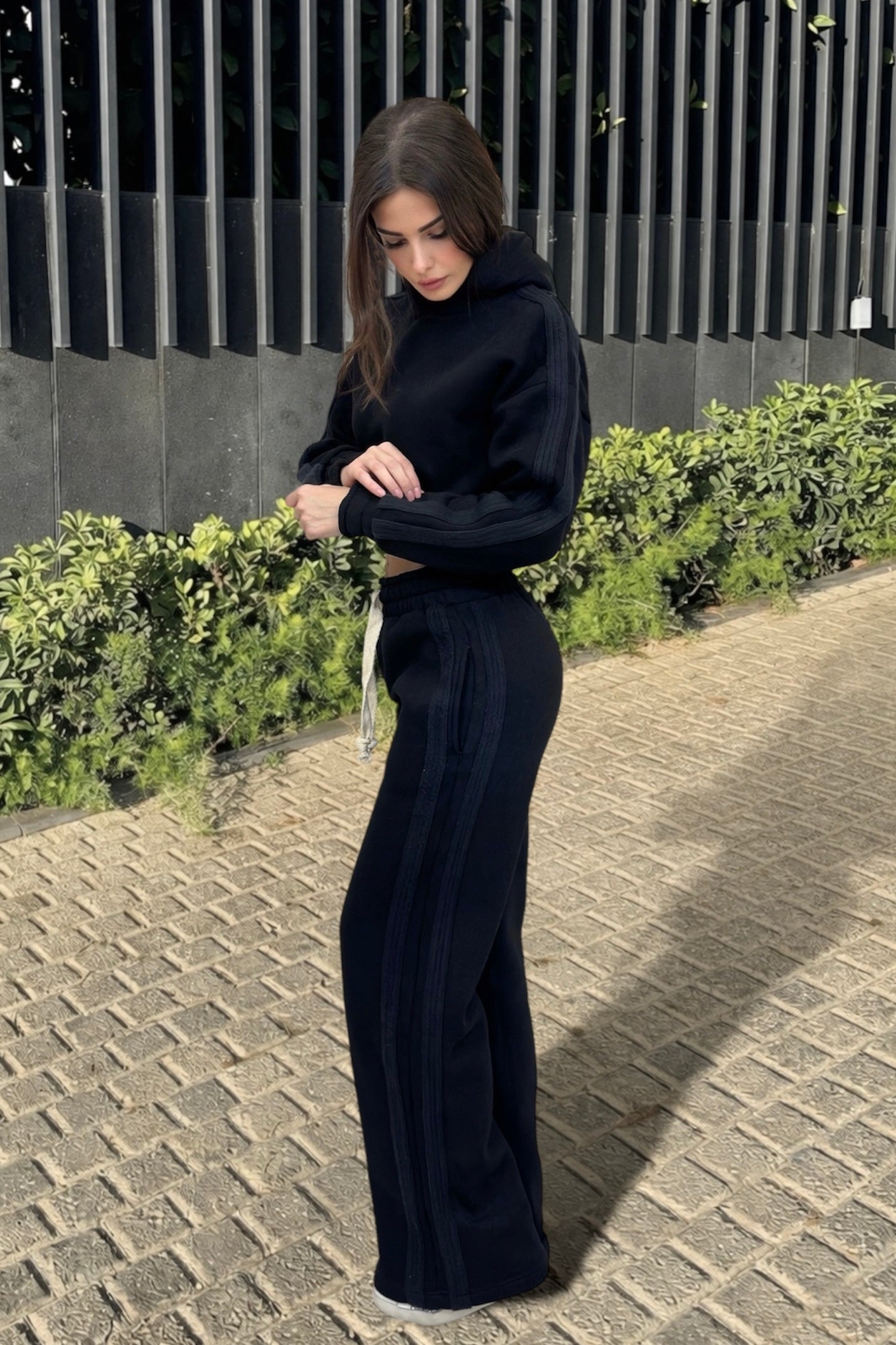 Woman in a black outfit standing on a sidewalk with greenery in the background