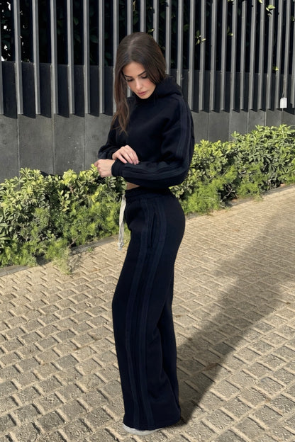 Woman in a black outfit standing on a sidewalk with greenery in the background