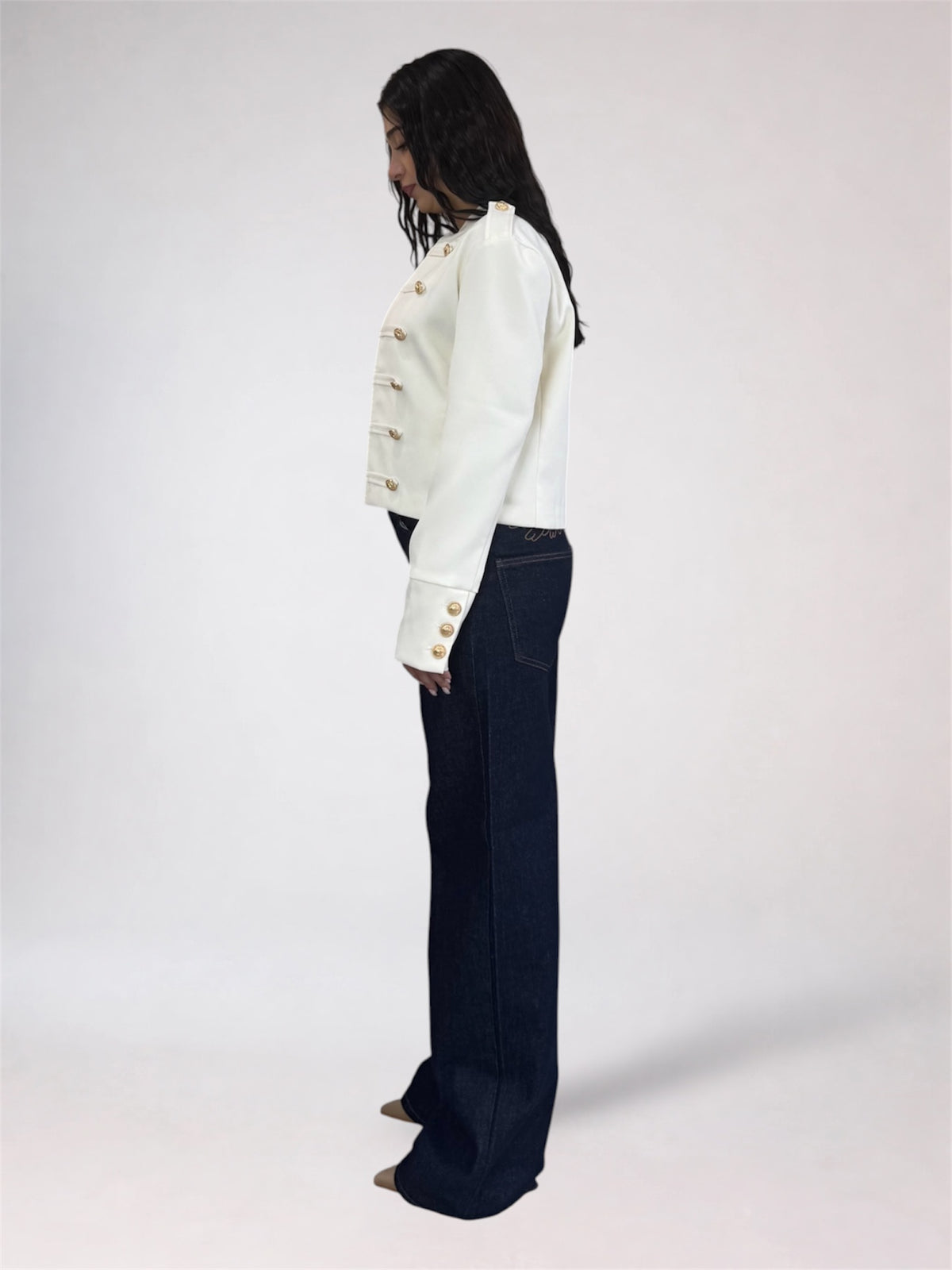 A model shown from the side profile wearing a white cropped jacket with gold buttons and dark navy wide-leg jeans, standing against a plain light gray studio background.