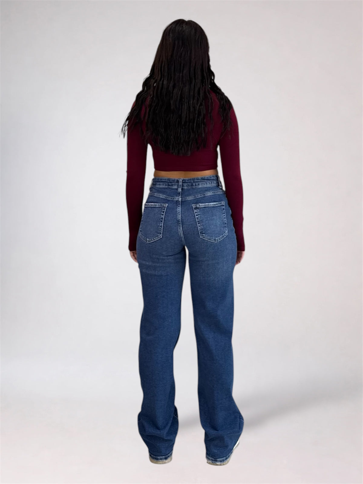 A woman viewed from behind wearing a dark burgundy long-sleeve crop top and high-waisted, wide-leg blue jeans against a plain light gray studio background.
