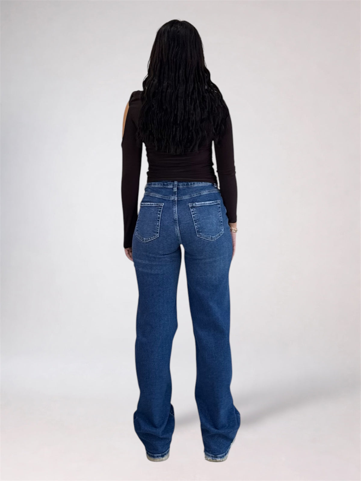 A woman viewed from behind wearing a black long-sleeve top and blue wide-leg jeans against a plain light gray studio background.