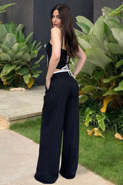 Woman in a black pinstripe jumpsuit standing outdoors with greenery around.