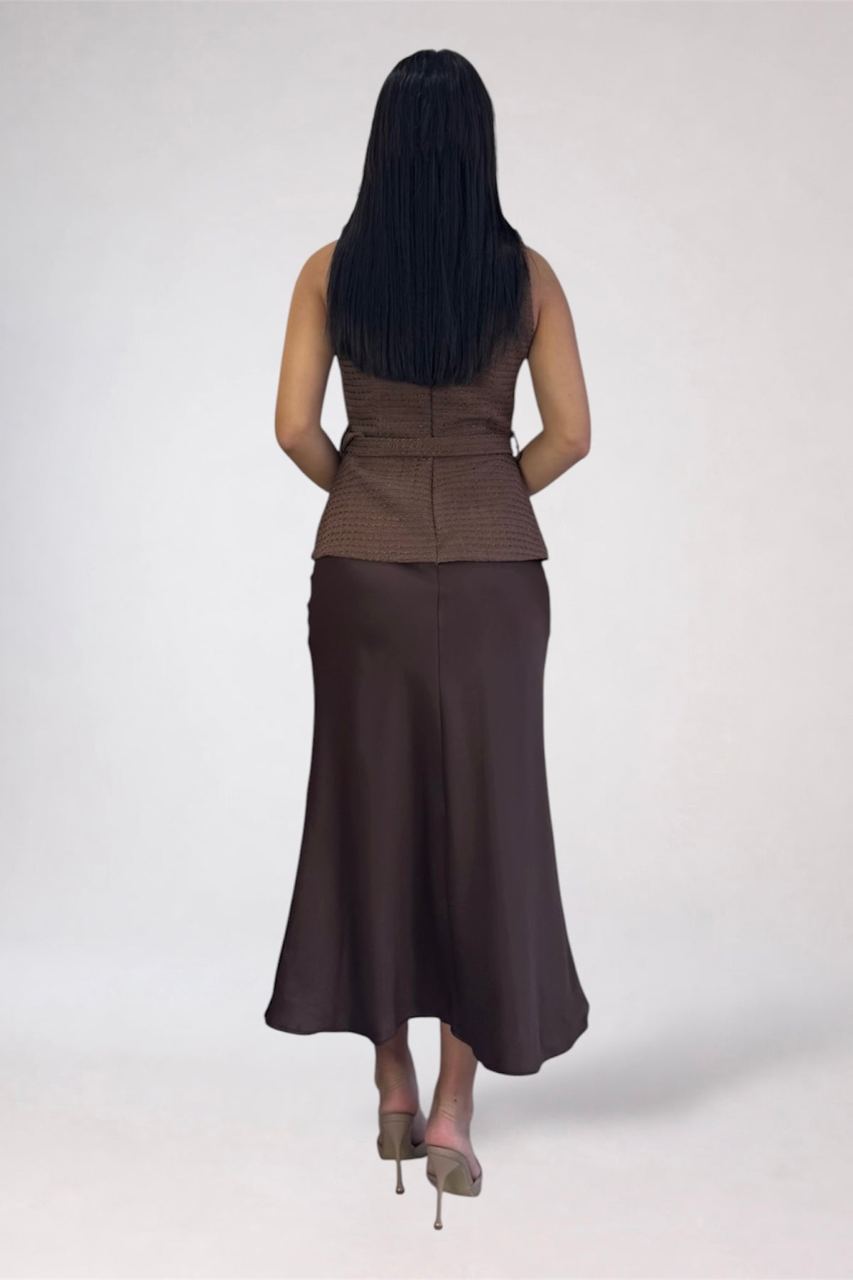 A woman shown from the back wearing a sleeveless brown textured top and a long dark brown skirt, standing against a plain light gray studio background.