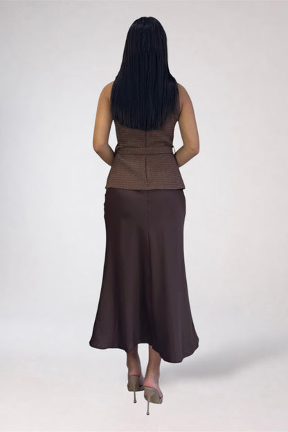 A woman shown from the back wearing a sleeveless brown textured top and a long dark brown skirt, standing against a plain light gray studio background.