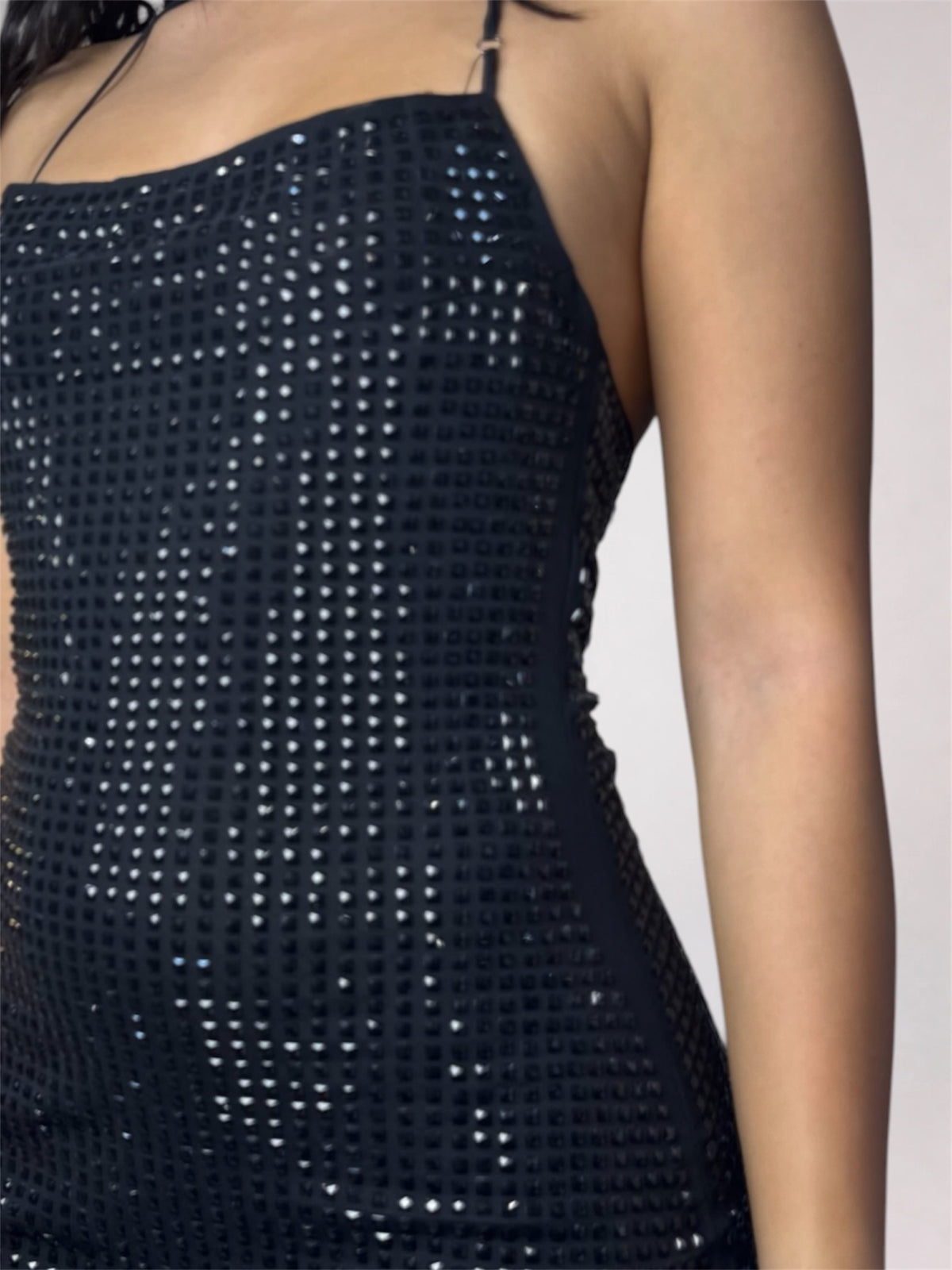 Close-up of a person wearing a black spaghetti-strap dress covered in small reflective rhinestones or sequins against a plain light background.
