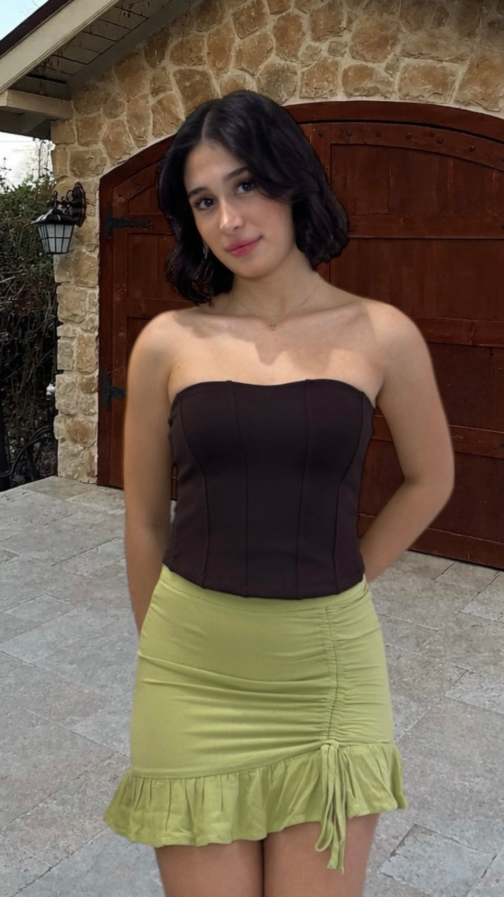 Woman wearing a brown strapless top and green skirt standing in front of a stone wall.