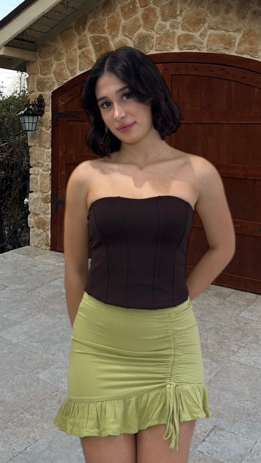 Woman wearing a brown strapless top and green skirt standing in front of a stone wall.