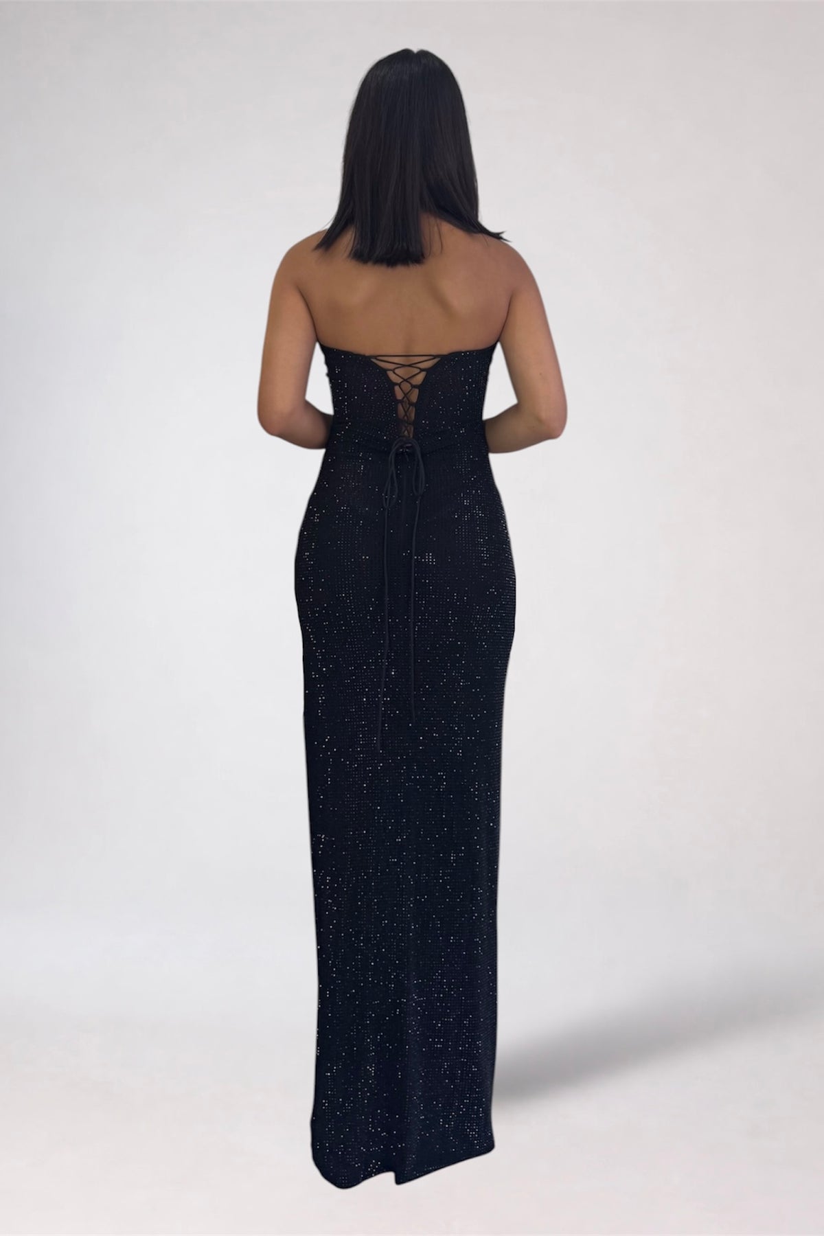 A woman viewed from the back wearing a strapless black evening gown with a lace-up open back and subtle shimmer, standing against a plain light gray studio background.