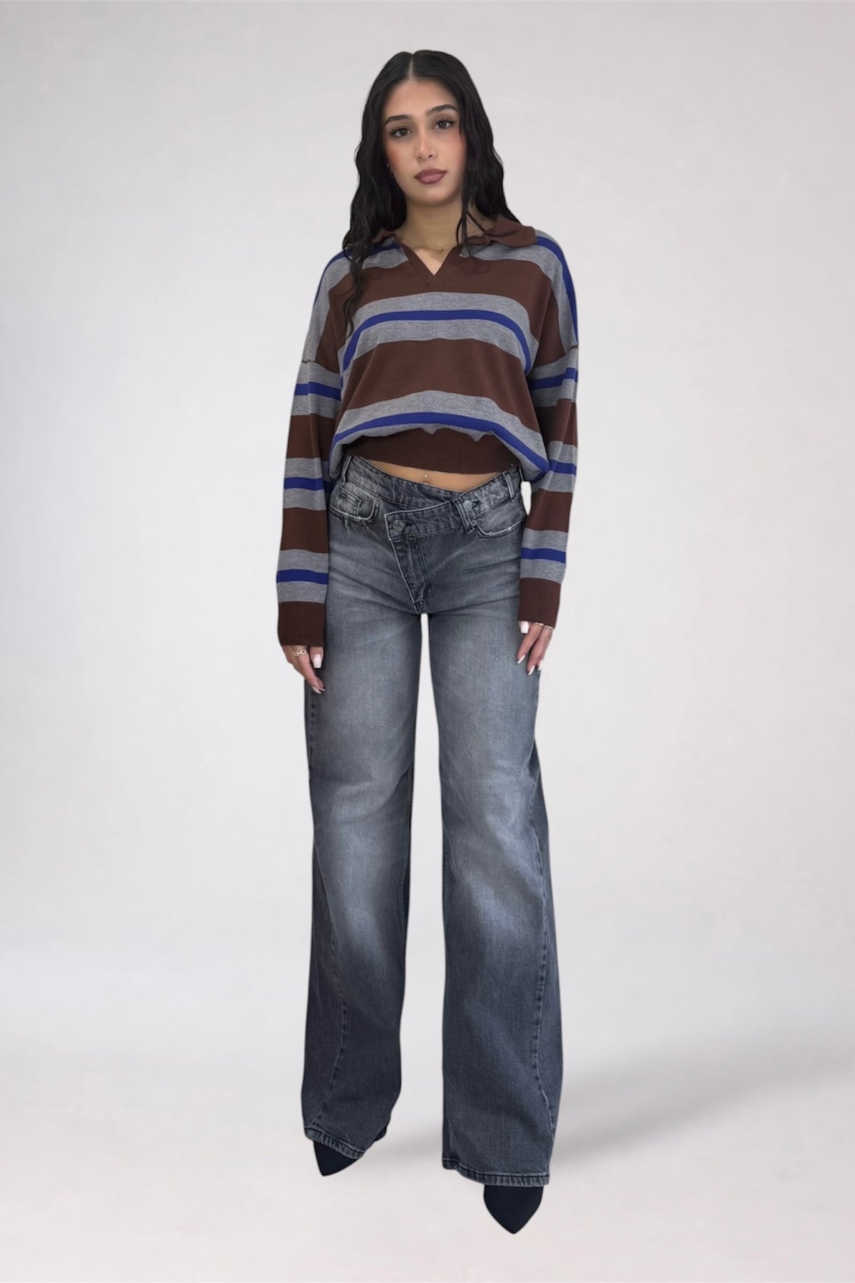 A model wearing a brown, blue, and gray striped long-sleeve crop top with wide-leg gray jeans, standing against a light gray studio background.