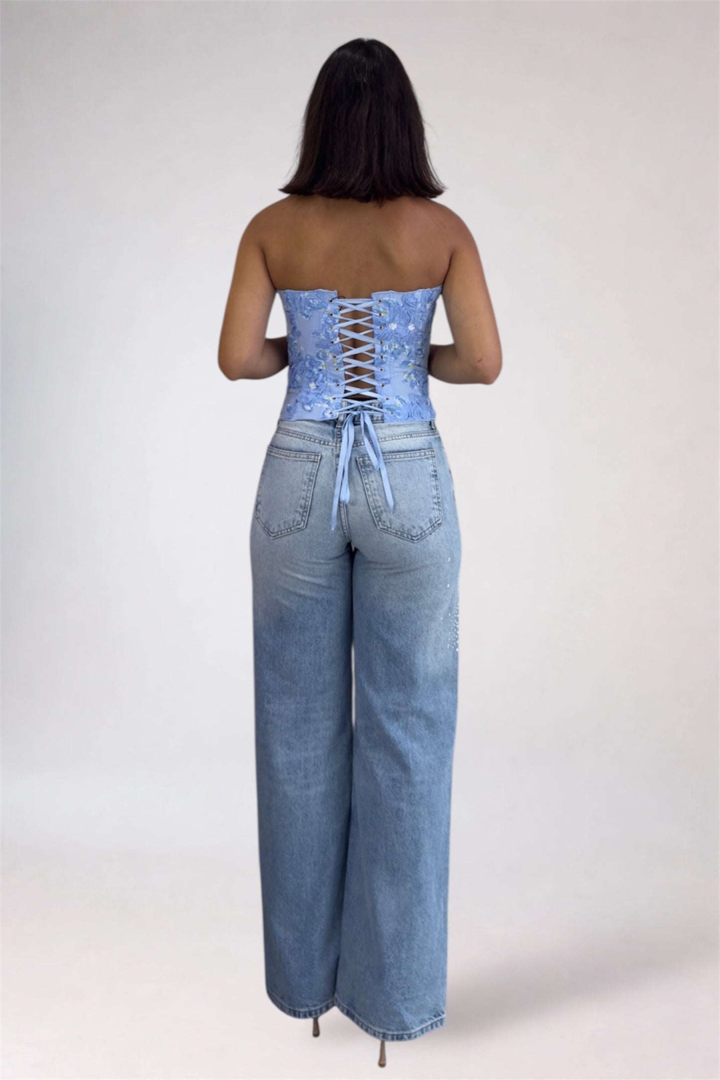 Woman wearing a strapless light blue top with lace-up back and high-waisted light blue jeans on a white background
