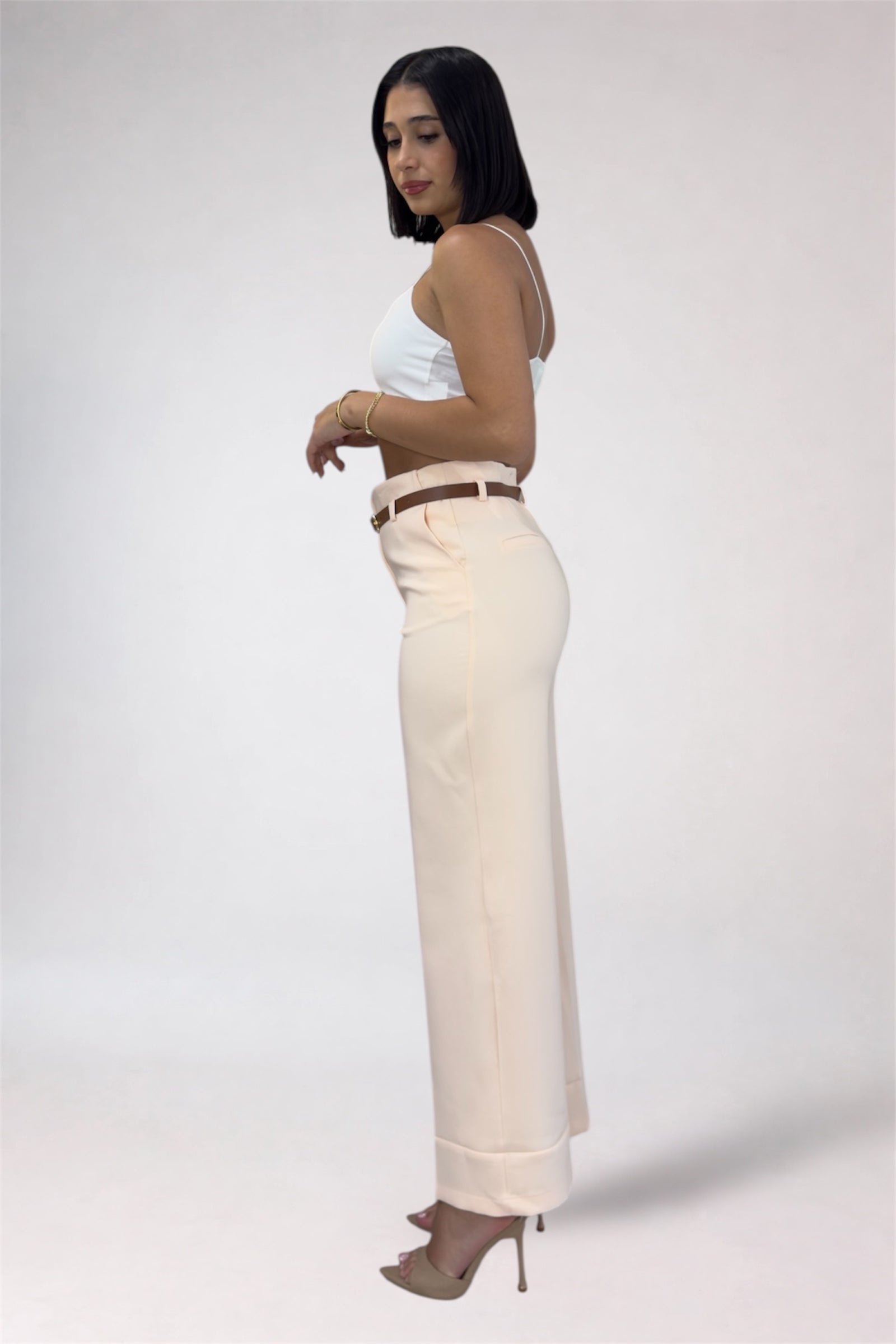 Woman wearing a white top and beige pants with a belt on a plain background