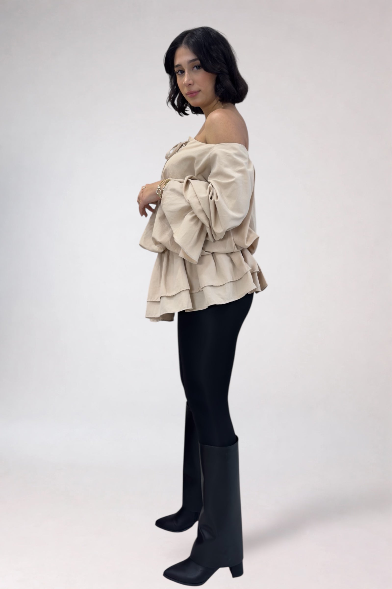 Woman wearing a beige off-shoulder ruffled top and black leggings with knee-high boots on a white background