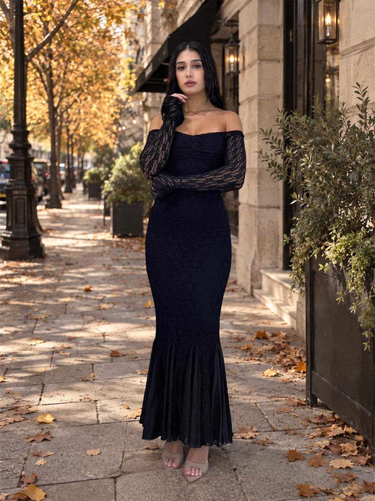 A woman stands on an outdoor city sidewalk wearing a fitted off-the-shoulder black dress with long sheer lace sleeves and a flared mermaid hem. The scene includes trees with yellow leaves, stone building walls, storefront awnings, and scattered fallen leaves.