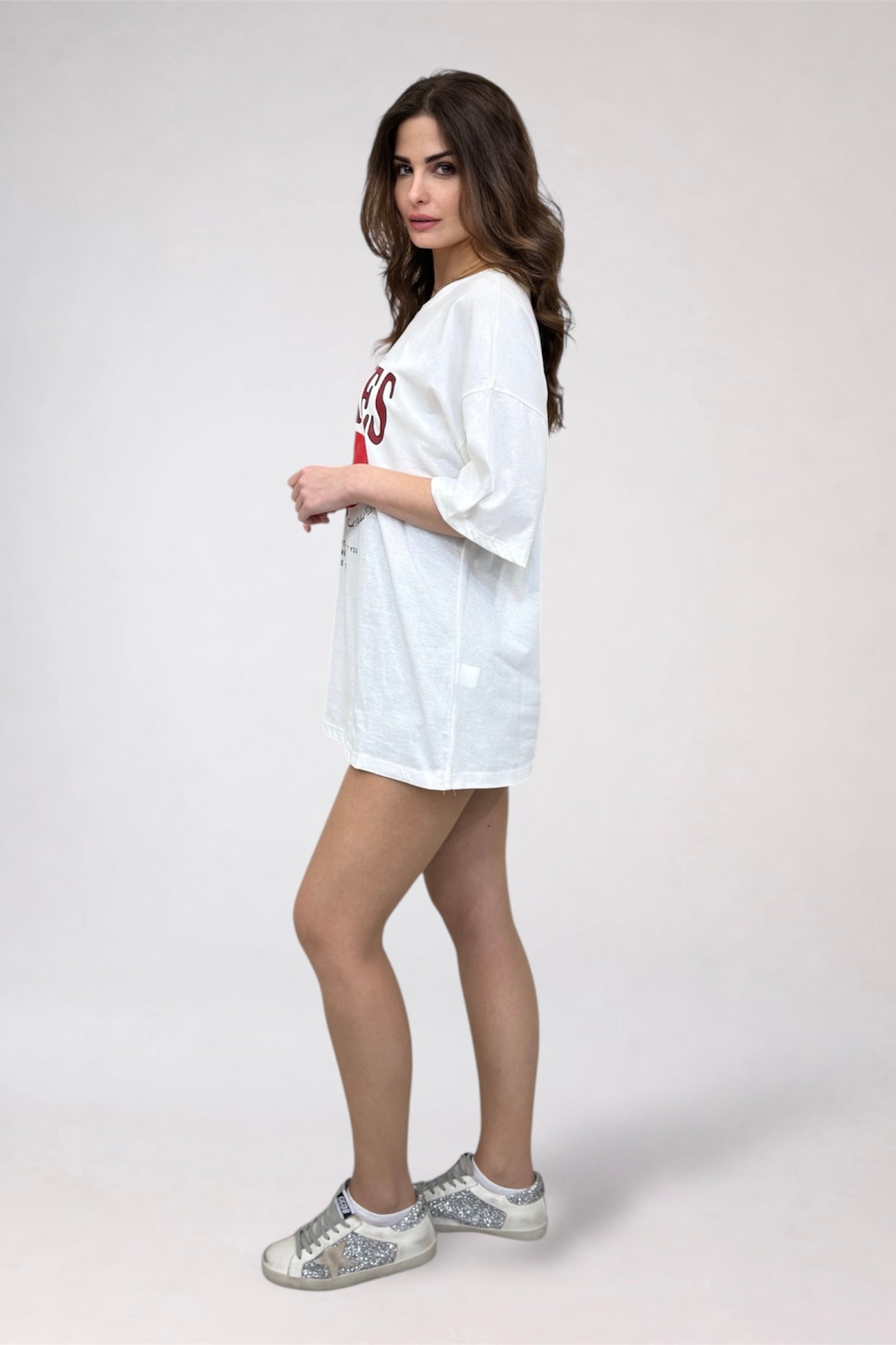 Woman wearing a white oversized sweatshirt with red text and silver glittery sneakers on a plain background