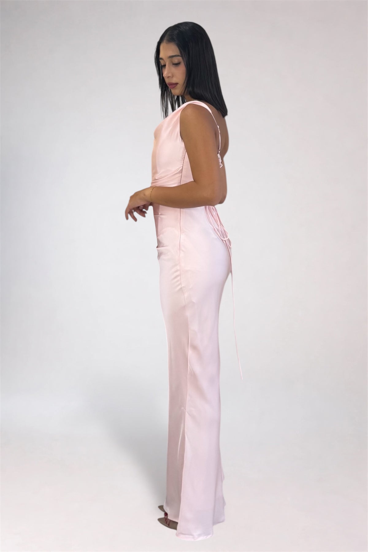 A woman modeling a light pink sleeveless maxi dress with a fitted bodice and flowing skirt, shown from the side against a plain light gray studio background.