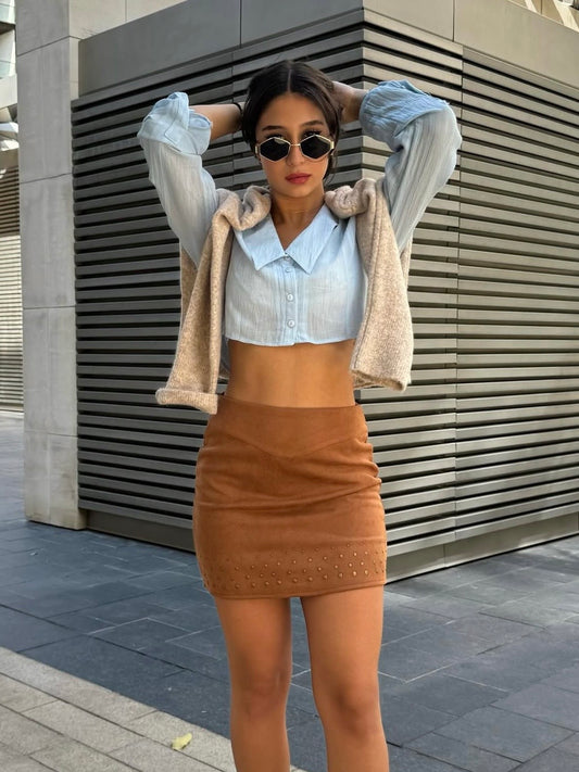 Woman posing in front of a modern building wearing a light jacket, white shirt, and brown skirt.