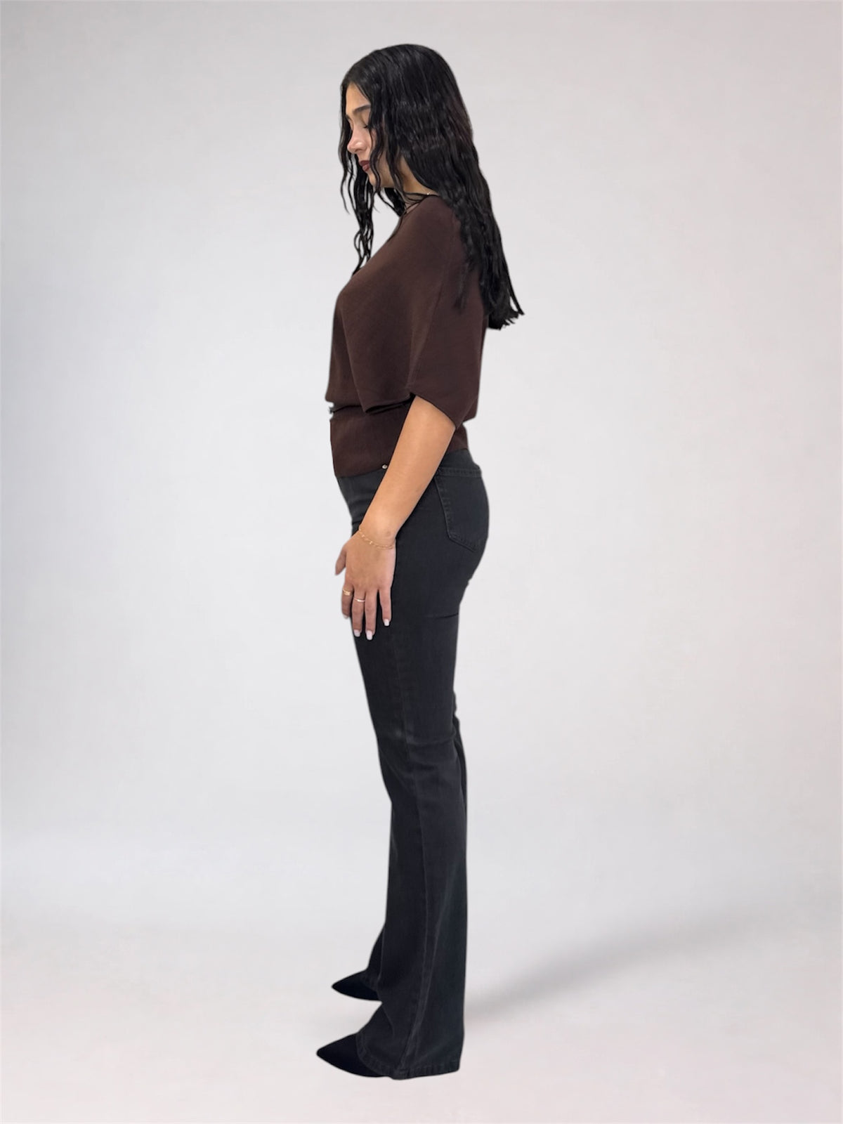 A woman shown in side profile wearing a dark brown short-sleeve top and black flared pants against a plain light gray studio background.