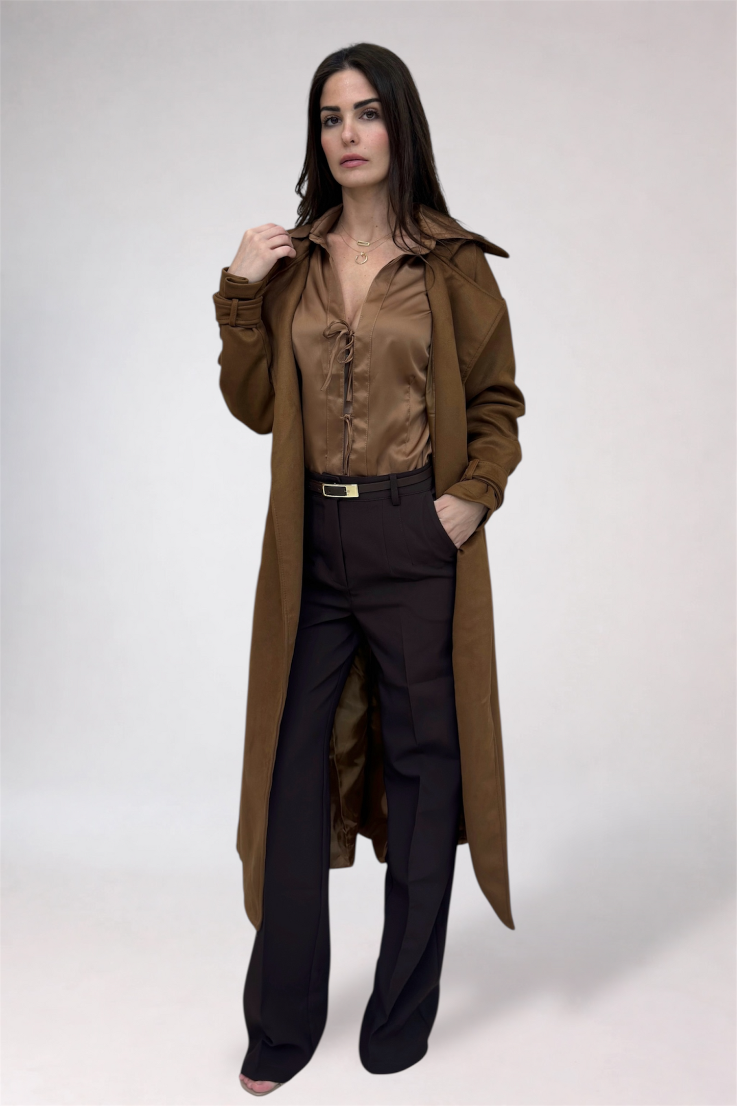 Woman wearing a long brown coat over a brown shirt and black pants on a white background