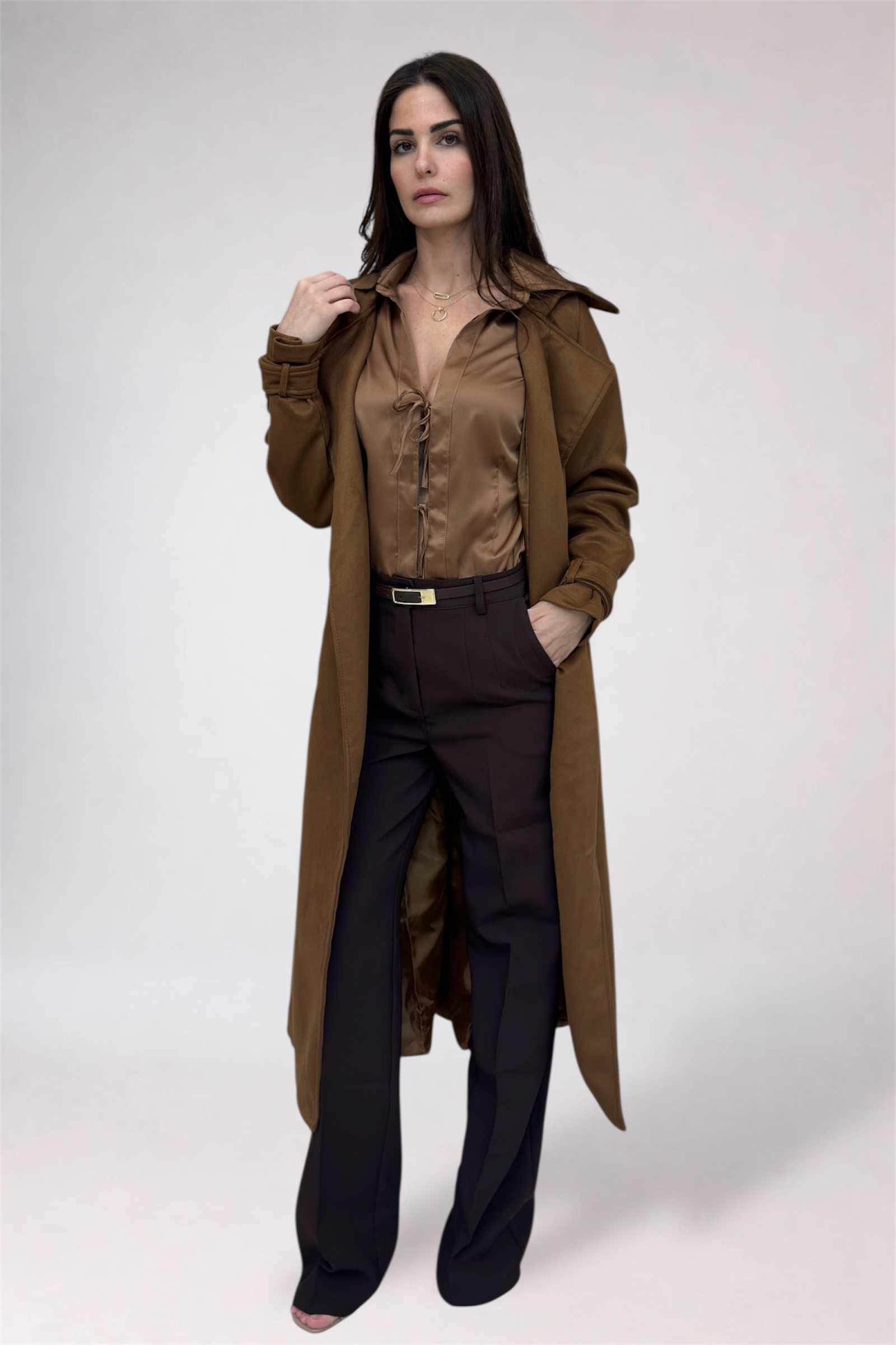 Woman wearing a long brown coat over a brown shirt and black pants on a white background