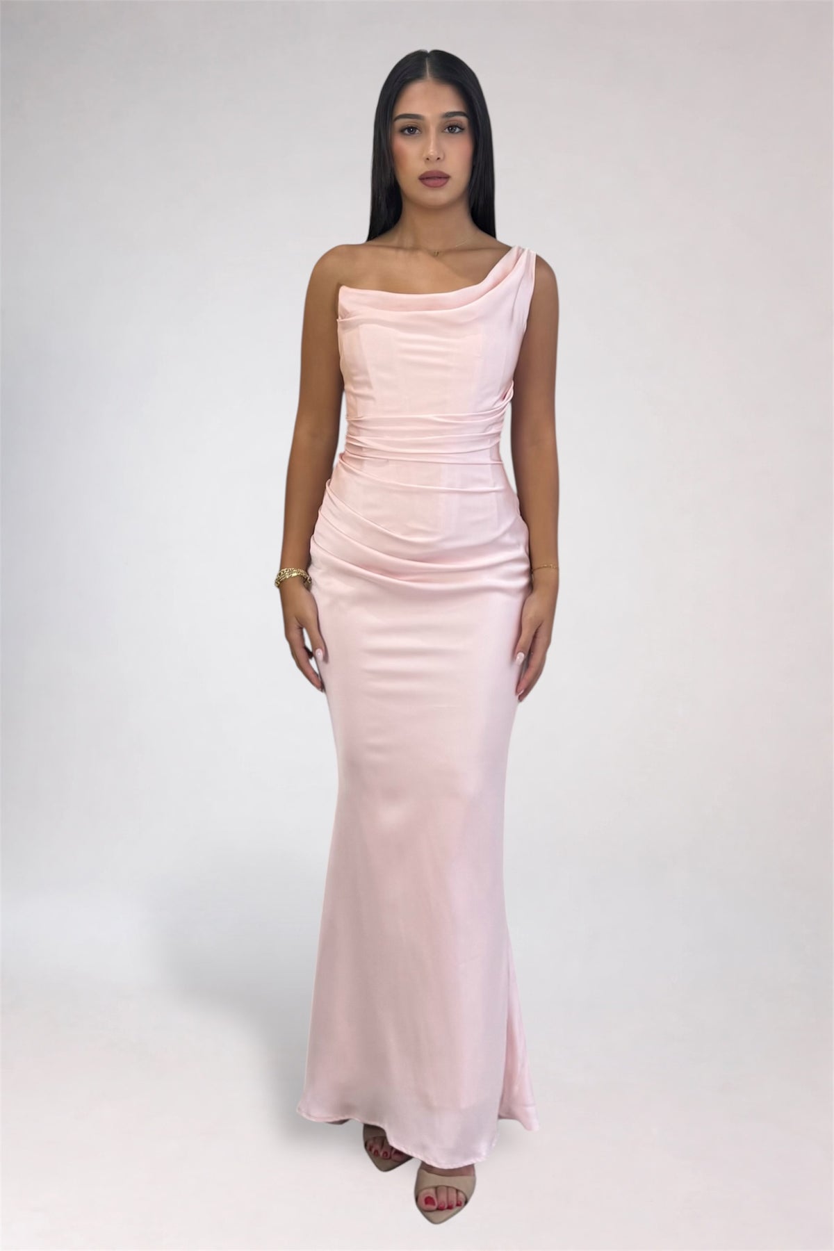 A woman modeling a light pink one-shoulder satin maxi dress against a plain light gray studio background.
