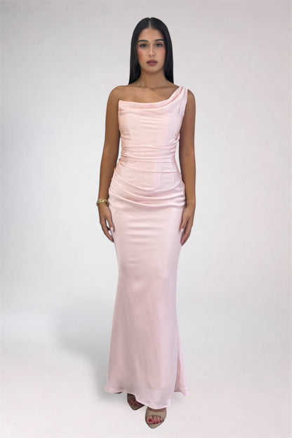 A woman modeling a light pink one-shoulder satin maxi dress against a plain light gray studio background.