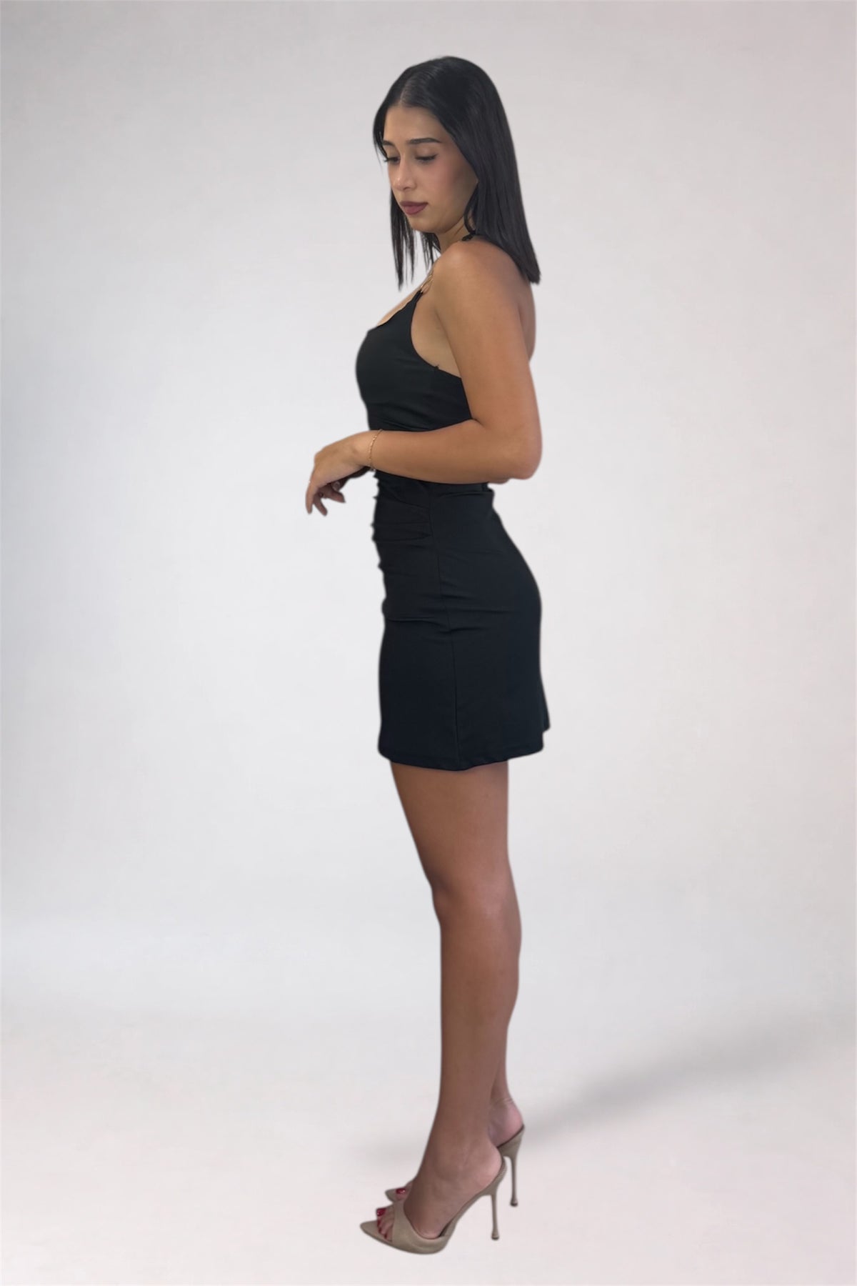 A woman modeling a black halter-neck mini dress with sheer nude high-heeled sandals, standing in profile against a plain light gray studio background.
