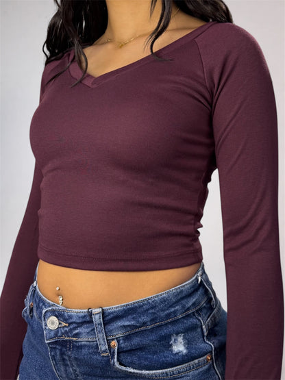 A woman shown in profile wearing a dark burgundy long-sleeve crop top, blue flare jeans, and off-white sneakers against a plain light gray studio background.