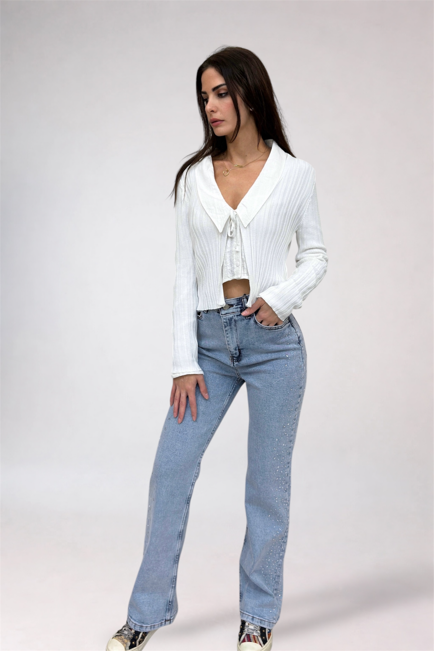 Woman wearing a white ribbed sweater and light blue jeans on a plain background