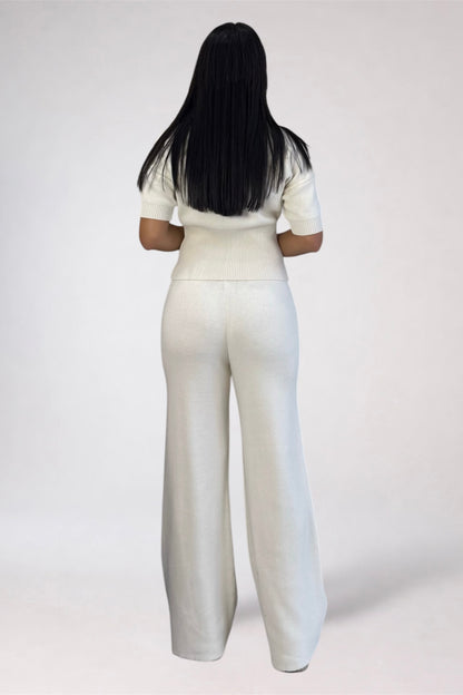 A woman viewed from behind wearing a matching cream-colored short-sleeve top and wide-leg trousers set, standing against a plain light gray studio background.