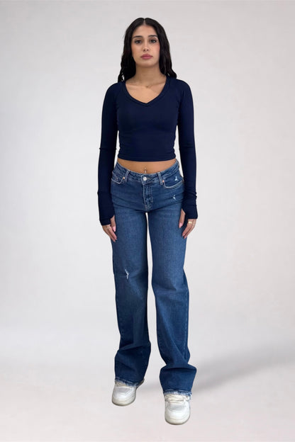 A woman modeling a dark navy blue long-sleeve crop top and blue jeans with white sneakers, standing against a light gray studio background.