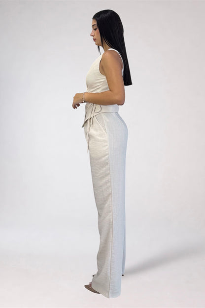 A woman stands in profile wearing a sleeveless cream top and matching cream wide-leg pants against a plain light gray studio background.