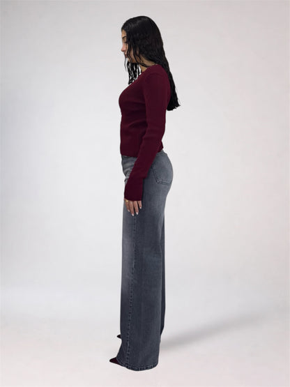 A woman shown in profile wearing a fitted burgundy long-sleeve top and high-waisted, wide-leg gray jeans against a plain light gray studio background.