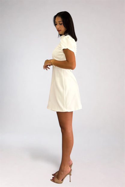 A woman modeling a short white dress with ruffled short sleeves and a high neckline, paired with nude high-heeled sandals, photographed against a light gray studio background.