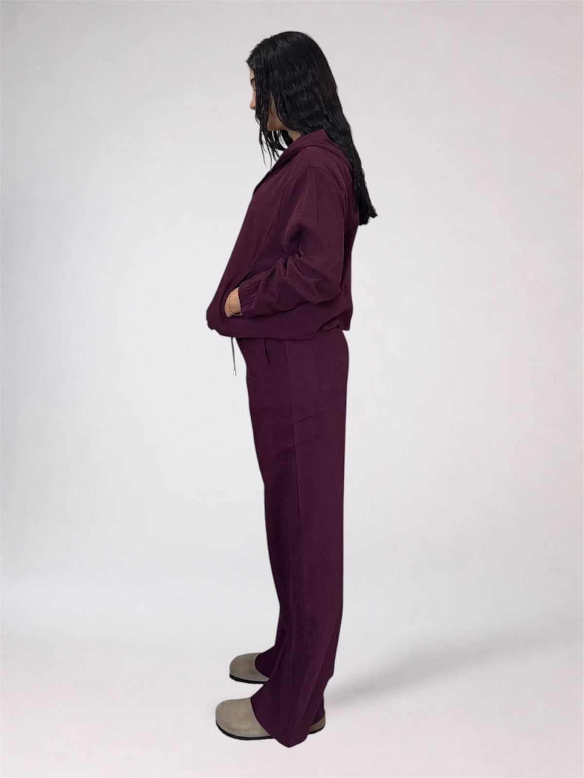 A model shown from the side profile wearing a matching deep plum purple lounge set consisting of a long-sleeve top and wide-leg pants, paired with gray slip-on shoes against a plain white studio background.