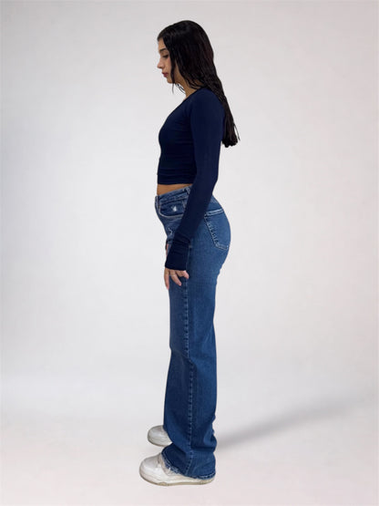 A woman shown in profile wearing a dark navy long-sleeve crop top, blue flare jeans, and white sneakers against a plain light gray studio background.