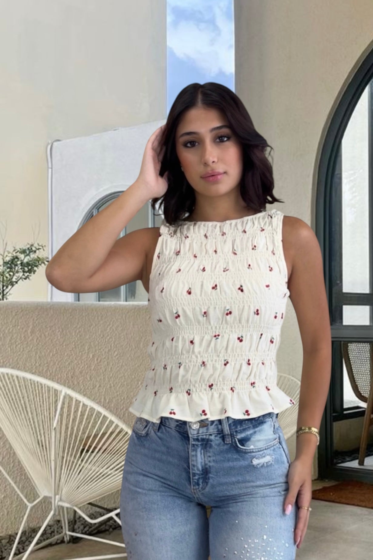 Woman wearing a white sleeveless top with red patterns and blue jeans indoors.
