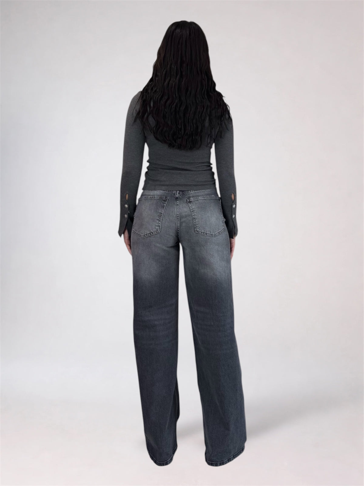 A woman viewed from behind wearing a dark gray long-sleeve top and wide-leg dark gray jeans against a plain light gray studio background.