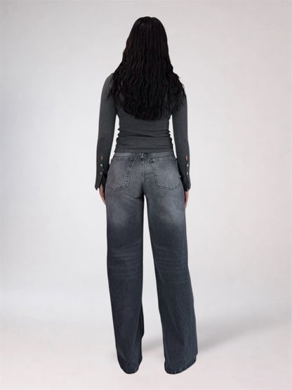 A woman viewed from behind wearing a dark gray long-sleeve top and wide-leg dark gray jeans against a plain light gray studio background.