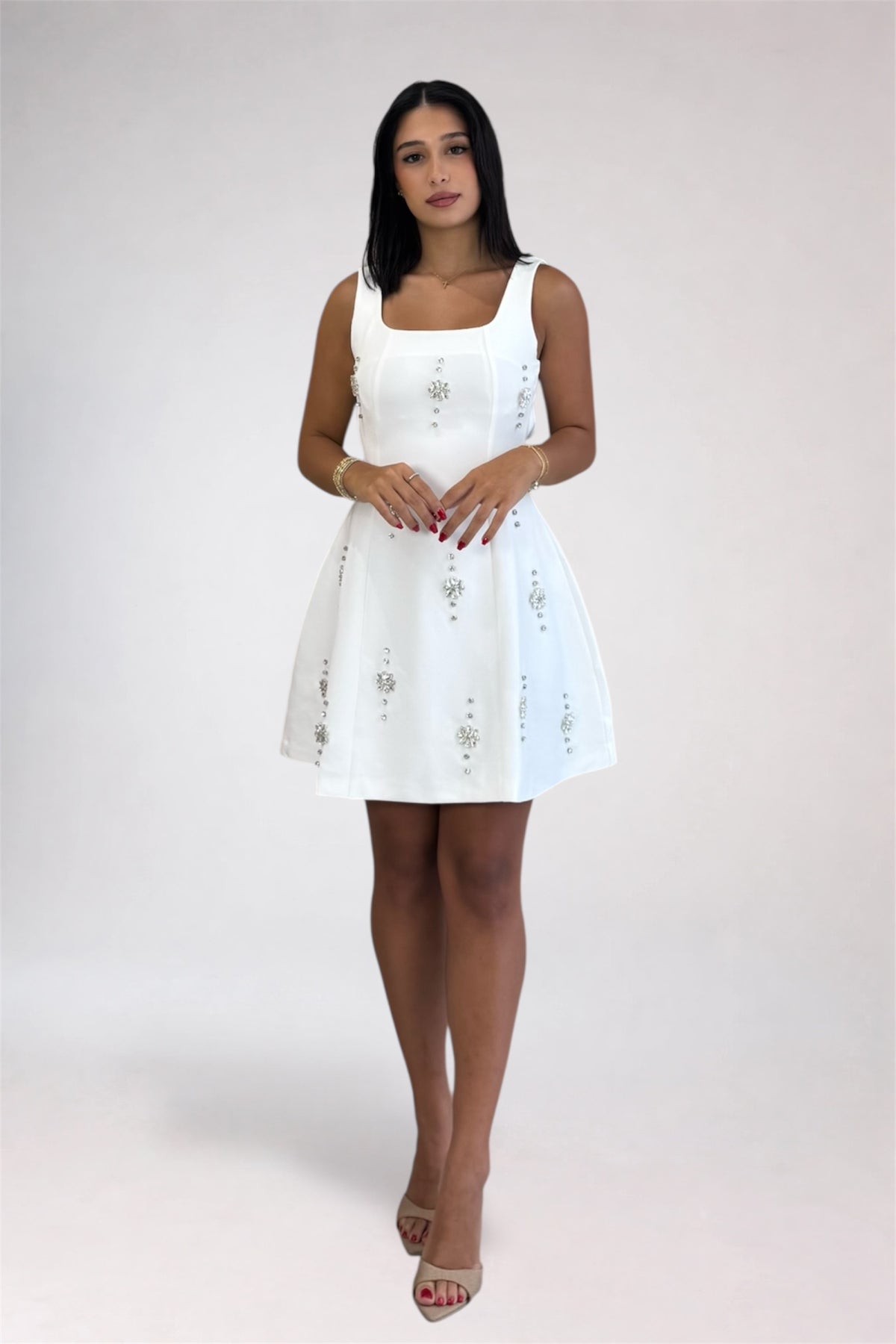A woman modeling a white sleeveless mini dress with embroidered details, standing against a light gray studio background.