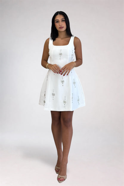 A woman modeling a white sleeveless mini dress with embroidered details, standing against a light gray studio background.